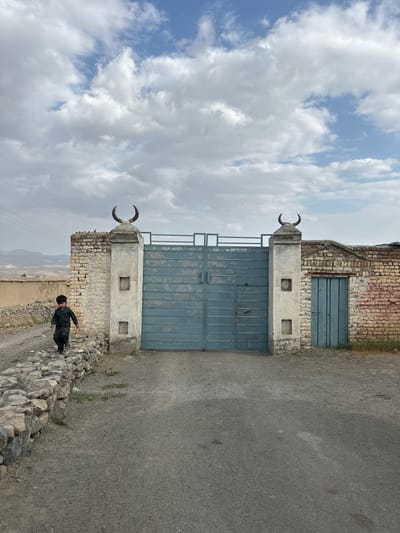 In the city of Kan Mehtarzai, Pakistan, a young boy wearing a dark green outfit walks along a stone-lined path next to a large blue metal gate. The gate is flanked by brick pillars, one of which has what appears to be animal horns mounted on top. To the right of the main gate, there is a smaller, blue wooden door set into a brick wall. The environment is dry and dusty, with some sparse vegetation growing along the edge of the property. In the background, there are hills and a cloudy sky.
