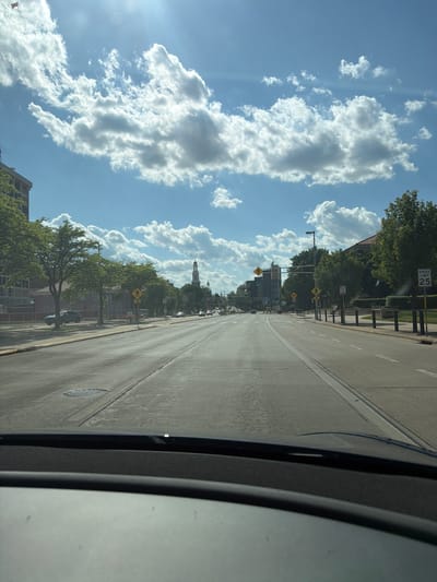 The image depicts a street in Madison, Wisconsin, United States, looking down towards the Wisconsin State Capitol building. The view is likely taken from inside a vehicle. The road is straight with marked lanes. Sidewalks line each side, with a mix of buildings and trees along the route. The sky is blue with large, puffy white clouds. Street signs and traffic lights are visible. A speed limit sign indicates 25 mph.