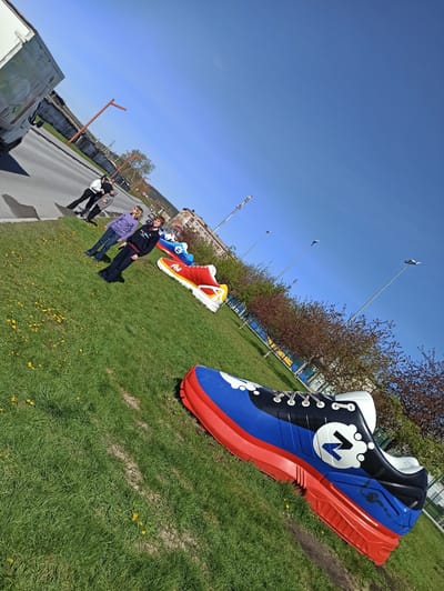 The image shows a row of oversized shoe sculptures on a grassy area alongside a road. The sculptures are painted in various colors, with the one closest to the camera being primarily blue, red, and black. Each shoe displays a different logo or design on its side.

Several people are standing near the sculptures further down the line. A vehicle, possibly a van, is partially visible on the left side of the road.

Trees line the grassy area behind the sculptures, and a building is visible in the background, suggesting an urban or suburban setting.

The scene is located in Satka, Russia, evidenced by the Russian flag colors on one of the shoe sculptures. The weather appears sunny.
