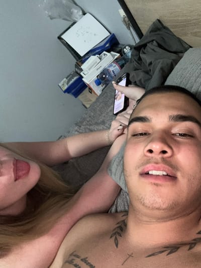 A selfie-style image shows a man lying on a bed with a woman. The man has short dark hair, brown eyes, and light facial hair. He has tattoos on his neck and chest. He is looking at the camera. The woman has blonde hair and is positioned below the man. She is not fully visible. Her lips are red. The man is holding a cell phone. Behind them, there are a water bottle and various boxes. The scene appears to be indoors, potentially in a bedroom. The location in the city of Blatets, Bulgaria is not discernible from the image.