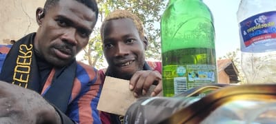 This image captures two men outdoors in Alayi, Nigeria. The individual on the left, a dark-complexioned male with a mustache, wears a black shirt, a blue and red plaid jacket, and a black scarf-like garment with yellow, reversed text "FEDEK2". He has a silver ear ornament in his left ear and looks directly at the camera with a neutral expression. The second individual, a dark-complexioned male with light blonde hair and a noticeable gap in his front teeth, wears a red patterned shirt. He smiles at the camera while holding a small, light brown piece of paper with handwritten text in his left hand.
In the background, a light-colored, weathered wall is visible on the left. On the right, a clear blue sky, green foliage from trees, and a portion of a building with a reddish-brown corrugated metal roof are present. In the mid-ground, to the right of the two men, a green glass bottle containing a light liquid is visible, displaying a partial label with a barcode and images. Next to it, a clear plastic water bottle with a reversed label "NKECELIJINYA ORAUMA" is also present, containing water. The foreground shows the top edge of a metallic, possibly gold-colored, container.