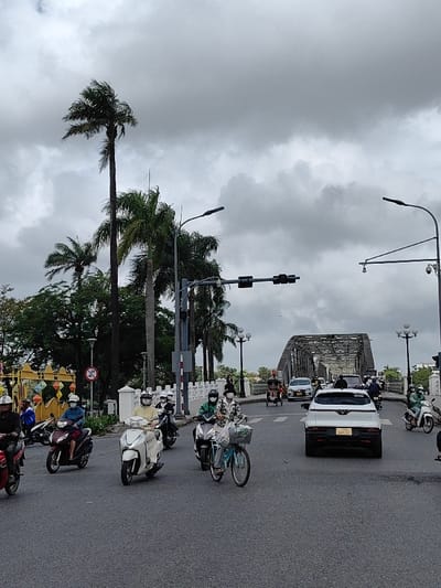 The image depicts an urban street scene on an overcast day, located in Thuận Hóa Ward, Huế, Vietnam, with the Trường Tiền Bridge visible in the background. The sky is uniformly gray with dense cloud cover.
In the foreground, an asphalt road occupies the lower third of the frame, showing signs of moisture. Numerous individuals are operating various vehicles. On the left, several motorbikes and scooters are present: a dark red scooter with a rider wearing a white helmet, a maroon scooter with a rider in a light blue jacket and white helmet, and a white scooter with a rider in a light yellow top and white helmet. Most riders are also wearing white or black face masks. A light blue bicycle with a front basket is visible in the mid-ground, ridden by a person in a patterned top, white helmet, and face mask, alongside a scooter.
In the mid-ground, a white VinFast VF 8 SUV is centrally positioned, traveling away from the viewer, displaying the license plate "75A-065.73". Traffic lights, currently unlit, are suspended over the road from a dark pole structure on the left. Multiple streetlights with dark, rectangular luminaires extend overhead on both sides of the road. A white concrete railing with ornate balusters lines the left side of the street, behind which numerous tall palm trees and dense green foliage are visible. A yellow and orange decorative structure with lantern-like elements is partially seen further left. A red "no entry" sign is also partially visible on the left. On the right, security cameras are mounted on a pole.
In the background, the metallic, arched truss structure of the Trường Tiền Bridge spans across the frame. Multiple vehicles, including cars, motorbikes, and a three-wheeled cyclo, are transiting the bridge. The bridge is also flanked by decorative streetlights similar in style to those on the foreground street.