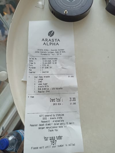The image shows a receipt from Arasta Alpha, located at Jalan Oversie Isdiman, Ruko W'dodo, Purwokerto Timur, 53114, Indonesia. The receipt is dated 13-04-2023 at 12:48, with a sales number of SAA01174452314383. The order was for one Kopi Susu Arasta (large, iced, less sugar, Arasta Milk, 50% Arabica : 50% Robusta, regular shot) and cost 31,000 Indonesian Rupiah. The customer's queue number is 167. The receipt also includes information about the WiFi network, which is powered by Starlink, with the SSID Arasta Alpha and password arastaalpha. It also states that orders can be picked up within 15 minutes, and to show this receipt. The receipt is lying on a black surface with a partial view of a blue liquid-filled container.