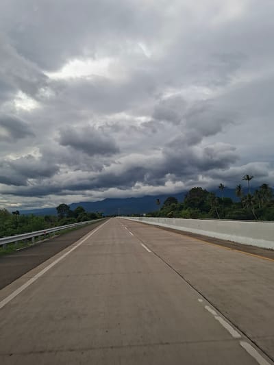 The image depicts a road in Baksukon, Indonesia, with a view of dense, ominous clouds overhead. The road itself is a grey concrete highway with a white line marking the edge. A barrier runs along the side of the road. Beyond the barrier is lush green vegetation, including trees and some palm trees. The sky is filled with dramatic, dark grey clouds, suggesting an impending storm.