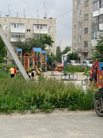 The image depicts a children's playground in front of apartment buildings, likely in Degtyarsk, Russia. A group of children and adults are present on the playground. In the foreground, a child in a black shirt and shorts holds a soccer ball, while another child in a yellow shirt stands nearby. Further back, a swing set with people on it is visible, along with a merry-go-round. Several cars are parked in the background, including a silver sedan and a darker colored car partially visible on the right. The playground equipment appears to be made of metal and plastic, with bright red, blue, and yellow colors. The surrounding area is overgrown with tall green grass and some trees. The buildings are multi-story concrete apartment blocks, typical of Soviet-era construction, with balconies on each level. The sky is overcast, suggesting a cloudy or potentially rainy day. The overall scene appears to be a typical residential setting on an ordinary day. There is no visible text in the image.