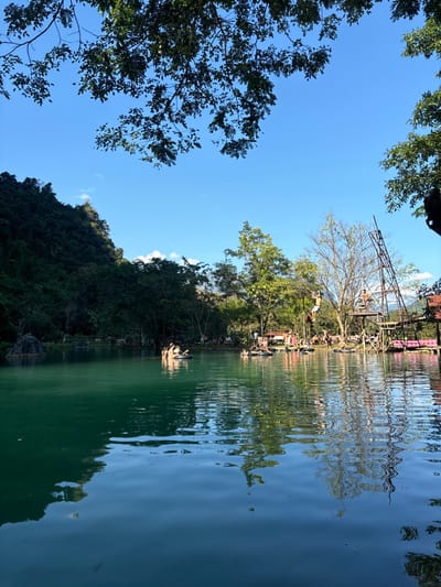 A verdant landscape meets a tranquil body of water under a bright, cloudless blue sky. Dense foliage lines the water's edge, contrasting with its reflective surface, mirroring the scene above. A tall structure stands amidst the trees, hinting at human activity and a destination for recreation. The surrounding natural beauty creates a serene atmosphere.