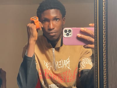 A male individual is captured in a mirror selfie taken indoors in Uturu, Nigeria. He is positioned centrally, facing the mirror and holding a light purple smartphone with a triple-lens camera array in his right hand to capture his reflection. His left hand is raised, holding an orange digital sports watch with a black face towards the viewer. The individual has dark skin, short, dark, curly hair, and is wearing a tan quarter-zip long-sleeve shirt with white and red graphic text designs on the chest and sleeves, layered over a dark long-sleeve undershirt. A portion of a dark-framed mirror with decorative gold patterns is visible on the right side of the frame. The background consists of a plain, light-colored wall.