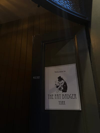 A sign for "The Fat Badger, York" is affixed to a dark, wooden door. The sign features a cartoon badger wearing a hat and scarf. Below the sign, small printed text reads "MIGHT/PORTER 07239412050". The door is positioned at an angle, and to the left is a glass window with a patterned design. The surrounding wall is dark with horizontal wood paneling. The overall lighting is dim.