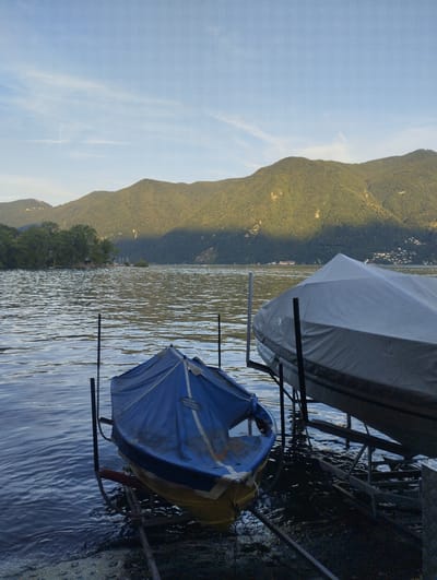 This image depicts a serene lakeside scene in Lugano, Switzerland, during what appears to be late afternoon or early evening, based on the golden light casting shadows. The foreground is dominated by two boats, both resting on submerged or partially submerged boat stands. The boat on the left is smaller and covered with a blue tarp, with some of its yellow hull visible. The boat on the right is larger and completely covered by a light gray tarp. Both boats are positioned on metal racks, indicating they are either being stored or are about to be launched.
The water of the lake, likely Lake Lugano, exhibits gentle ripples, reflecting the soft, diffused light. The surface of the water is a deep blue-gray, with lighter patches where the light hits. Along the shore to the left, there's a small area of vegetation and debris.
In the mid-ground, the lake stretches out, meeting a verdant, tree-covered shoreline. A cluster of trees on the left shore adds depth to the scene, and further back, several sailboats are visible, moored or sailing on the water.
The background is defined by the majestic presence of mountains, densely covered in lush green vegetation. These mountains are partially illuminated by the setting sun, creating a stark contrast between the sunlit slopes and the deep shadows cast across them. The sky above is a clear, pale blue with a few wispy white clouds and contrails, suggesting fair weather. The overall atmosphere is one of tranquility and natural beauty.