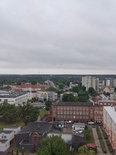 This is a vertical, high-angle shot of an overcast cityscape, likely taken from a tall building. The scene is dominated by the grey sky and a dense arrangement of residential and commercial buildings. Trees and greenery are interspersed throughout the urban landscape, particularly along the sides of roads and in smaller green spaces.
Several multi-story buildings are visible, characterized by their varied architectural styles and colors. One prominent building on the right is a tall, light-colored apartment building with a distinctive pattern of orange and white on its facade. Adjacent to it is a darker brick building with many uniformly spaced windows. Other buildings in the scene range from modern, white structures to older, multi-story blocks in shades of grey, yellow, and red.
A street runs through the middle ground of the image, with several parked cars visible. The presence of cars, sidewalks, and road markings suggests a typical urban environment. A telecommunications tower is visible in the distance, silhouetted against the hazy horizon.
The weather appears to be overcast, creating a diffused light and a slightly muted color palette across the scene. The time of day is difficult to determine precisely due to the lack of strong shadows, but it appears to be daytime.
No people are clearly visible in the image. The overall impression is one of a lived-in urban environment under a cloudy sky. There is no visible text in the image.