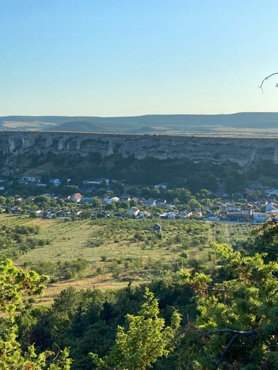 This is a vertical, eye-level, outdoor, long-shot photograph capturing a pastoral landscape at the base of a sheer cliff face. The sky is a clear, light blue with no visible clouds. In the middle ground, a village is nestled amongst lush green trees. The village consists of numerous residential buildings with red, grey, and white roofs, interspersed with more vegetation. The buildings appear to be of varying sizes and architectural styles, suggesting a well-established community.
The foreground is dominated by the branches and foliage of evergreen trees, some of which are in sharp focus, while others recede into a soft blur. These trees frame the view of the village and the distant landscape.
Beyond the village, a prominent, light-colored cliff face rises dramatically. The cliff has a textured, rocky surface with visible striations and formations. Above the cliff, rolling hills stretch into the hazy distance, their surfaces appearing as a patchwork of light brown and green, indicating fields or drier vegetation. The overall impression is one of a serene, rural setting bathed in natural light, likely during the daytime. The location is specified as Krasnomakskyi rural settlement, Ukraine.