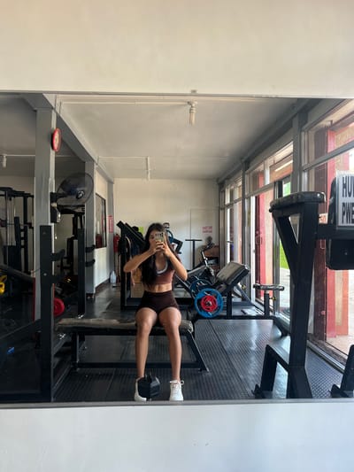 In the city of Cuyo, Philippines, a young woman is captured in a full-length mirror selfie within a gym. She is wearing a brown sports bra and brown shorts, with white sneakers. The gym appears to be equipped with various weightlifting machines and benches. A black oscillating fan is mounted on a gray pillar to the left, and a red-faced clock is visible on the same pillar. Large windows line the right side of the gym, revealing an outdoor view. The floor is covered in black textured matting. The woman is holding a smartphone in both hands, taking the photograph.