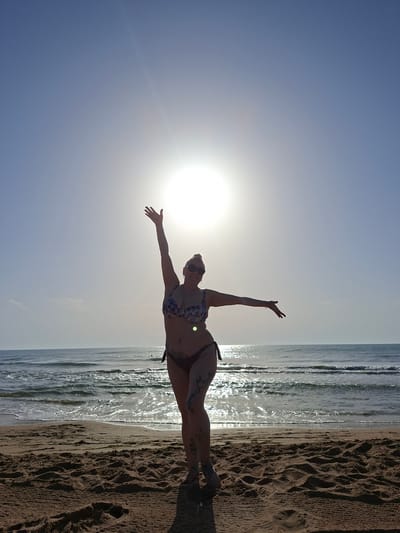 A woman wearing a bikini stands on a sandy beach in La Mata, Spain, with her arms outstretched towards the sun. The sun is visible in the upper center of the frame, partially obscured by a slight haze. The ocean is behind her, with gentle waves rolling onto the shore. The sand is wet near the water's edge and dry further up the beach, with visible footprints and sand textures. The woman has tattoos on her legs and is wearing sunglasses. The sky is a clear blue, transitioning to a lighter shade near the horizon where the sun is positioned.