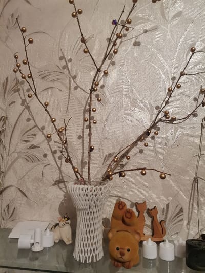 This image captures a close-up indoor scene featuring a decorative arrangement on a clear, reflective surface, set against a light-colored, textured wallpaper.
At the center, a vase holds several bare branches. The vase itself is covered with a white, open-weave, mesh-like material, giving it a distinct patterned appearance. The branches are adorned with numerous small, spherical, golden-colored beads, resembling berries or pearls, giving them a festive and ornamental quality. A few small, darker, possibly purple or brown, decorative elements are also scattered among the golden beads on the branches. The branches cast prominent, slightly blurred shadows on the wallpaper behind them.
The wallpaper features a subtle, repeating embossed or textured pattern of swirling leaves and floral motifs in a light, possibly off-white or cream hue.
Arranged on the clear, reflective surface in front of the vase are several small decorative items. From left to right:
* Two small, white, cylindrical objects resembling flameless LED candles, accompanied by a small white card or folded paper.
* A small ceramic figurine of a cat with white fur, black patches, and orange markings on its face, depicted with an expression that appears grumpy or startled.
* To the right of the central vase, there is a fuzzy, light brown teddy bear figurine, possibly a coin bank, lying on its back with its paws pointing upwards.
* Behind the teddy bear are two wooden silhouette figurines of cats, one slightly taller than the other, standing upright on a small base.
* Further to the right, three more small, white, cylindrical objects, identical to the ones on the far left, are visible.
* On the far right edge of the frame, a dark, cylindrical object (possibly a container or holder) is partially visible, and a white wire or cord hangs down the side, partially obscured by the branches.
The objects are placed on a transparent surface, likely glass, which shows faint reflections of the items and the light above. The overall lighting suggests an indoor setting, possibly with artificial illumination, creating a warm and decorative ambiance. There is no visible text in the image.