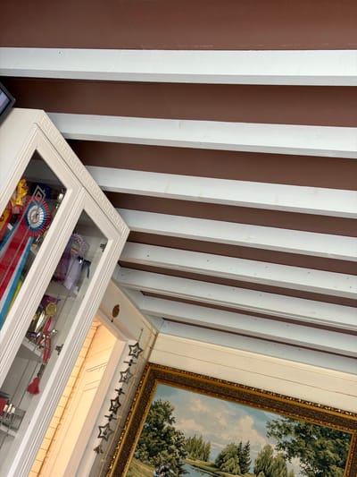 The image shows a close-up, high-angle view of a room interior, focusing on the ceiling and a display cabinet filled with trophies and ribbons. The ceiling features a dark brown finish with evenly spaced white beams. To the left, a white cabinet with a glass door displays multiple awards, including ribbons in red, white, and blue, and various trophies. The cabinet is adorned with a decorative Greek key pattern around its top edge. Below the cabinet, a glimpse of a wall is visible, leading to a doorway. To the right of the doorway, a large framed landscape painting hangs on the wall. The painting depicts a serene scene with trees and a body of water. A string of star-shaped ornaments with small bells hangs from the wall next to the doorway. The overall impression is of a well-decorated space, possibly a home, with a focus on achievements. The location of Podzhigorodovo, Russia is not discernible from the visual information in the image.