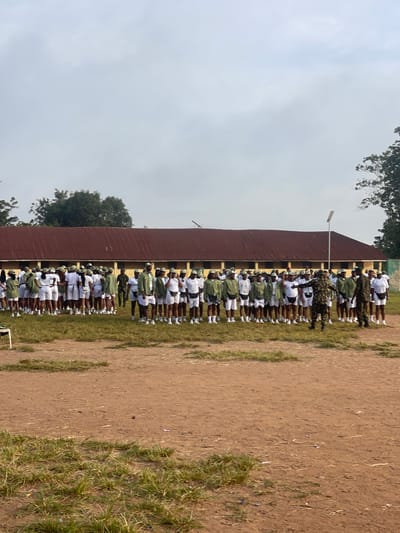 The image captures a group of individuals, likely uniformed cadets or participants, standing in formation on a grassy area in front of a long, yellow building with a corrugated iron roof. The majority of the group are dressed in white shorts and shirts with olive green vests and caps, suggesting a uniform. Several individuals in the foreground are wearing full olive green camouflage uniforms and appear to be instructors or supervisors, one of whom is in motion, possibly demonstrating a drill. The environment appears to be an outdoor training or assembly ground, with a dirt path visible in the foreground and trees in the background. The sky is overcast. Based on the context of similar images from this location, this scene is likely at the NYSC Orientation Camp in Ise - Ekiti, Nigeria, a common site for national youth service orientation programs.