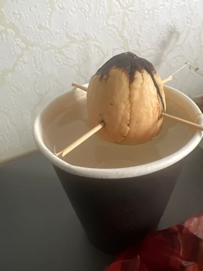 An avocado pit, suspended by three toothpicks, is submerged in water within a dark brown paper cup. The pit has a dark, possibly chocolate, residue on its upper portion. The cup sits on a dark, flat surface, with a textured, pale wall in the background. The scene is indoors and appears to be a home setting. The image does not contain any recognizable landmarks or elements that would indicate its specific location within the city of Pskov, Russia.