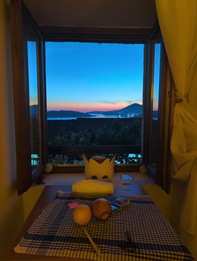 The image, oriented correctly, depicts an indoor scene looking out a window during twilight. Outside, the sky displays a striking gradient, transitioning from a deep blue at the top to a vibrant band of orange and pink along the horizon, indicative of either sunrise or sunset. Below the colorful sky, a dark mountain range stretches across the horizon, possibly bordering a body of water, with faint lights visible on the distant land. In the immediate exterior foreground, dark rooftops with terracotta tiles are visible, nestled among lush green trees and foliage.
Inside, the view is framed by a dark wooden window frame, with a reflection of the outside sky visible on the lower pane. Light-colored (likely yellow or beige) fabric curtains are draped above the window. To the right of the window, a wooden table is covered with a blue and white checkered tablecloth. On the table, several objects are arranged: a yellow, crown-like object with red detailing, resembling the "Super Crown" from the Mario series; a cylindrical yellow or cream-colored object (possibly a candle or small vase); a reddish-brown apple; and a yellowish-orange fruit, possibly an orange or tangerine. A small pink sticky note is tucked beneath the orange, and a dark pen or stick lies on the tablecloth near the apple. Two small, round balls, one yellow and one red, are partially visible on the window sill or table edge. The indoor lighting is warm and soft, casting a cozy ambiance that contrasts with the cool blues and vibrant pinks of the natural light outside. No explicit activity is taking place, but the scene conveys a tranquil moment of observation. The location context for this scene is Podličak, Montenegro.