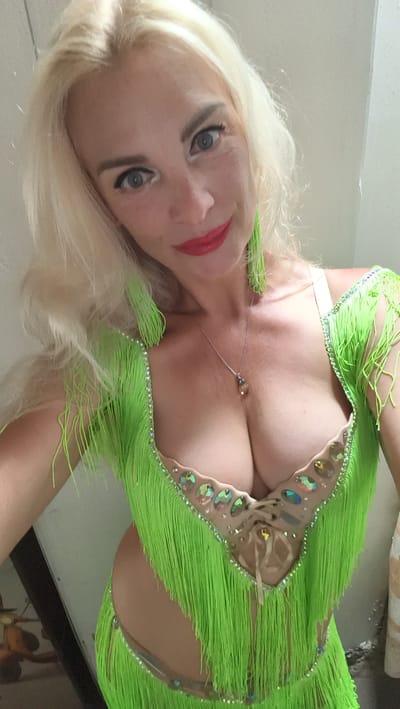 The image is a selfie taken by a blonde woman wearing a neon green, fringed costume adorned with rhinestones. She has red lipstick and is looking at the camera. The background appears to be an indoor setting with a plain, light-colored wall and some out-of-focus objects in the lower left corner, suggesting it is a private residence or a dressing room. There is no visible indication of the city of Kuşadası, Turkey.