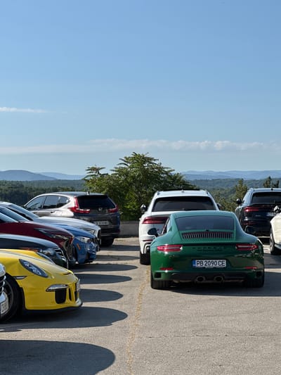 The image depicts a parking lot with multiple vehicles, situated in Arbanassi, Bulgaria, with a scenic mountainous landscape in the background under a clear blue sky. The foreground is dominated by several cars parked in a line. A prominent green Porsche 911 Carrera is in the center, displaying a Bulgarian license plate "PB 2090 CE". To its left, a yellow sports car is partially visible. Behind the Porsche, a white SUV and a black SUV are parked. Further to the left, a silver Honda CR-V SUV with a Bulgarian license plate "CB 3030 MX" is visible, along with other cars. The terrain is paved, and shadows from the vehicles are cast on the ground. Trees and bushes are present, and a low wall runs along the right side of the parking area. The overall scene suggests a gathering of vehicles, possibly at a scenic overlook or a car event.