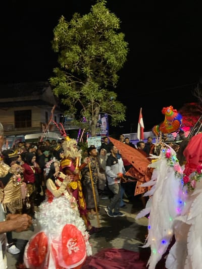 A nighttime street scene in Kedungbanteng, Indonesia, depicts a procession. The environment is illuminated by artificial light against a dark sky. In the background, a two-story building with a dark roof and window is visible on the left. A tall, leafy tree stands centrally, with a banner displaying text and images attached to its lower trunk, adorned with multi-colored string lights.

A dense crowd of individuals, primarily adults, lines the street, observing the procession. On the far right, an Indonesian flag (red and white) is visible on a pole, alongside a large, inflatable, multi-colored rooster balloon.

In the foreground and midground, several costumed figures move through the scene. On the central-left, a figure wears a gold-toned, ornate costume. Adjacent to this, a prominent figure is dressed in an elaborate monkey-like costume featuring a light-colored, shaggy head with visible teeth/tusks and what appears to be red and gold armor-like segments. This figure holds a long, dark staff. Beside the monkey-like figure, another person wears a voluminous white gown with multiple ruffled layers and a light-colored top, holding a small, white object to their mouth. Partially visible in the lower foreground are two circular, red objects. On the central-right, an individual is equipped with large, white, textured wings adorned with multi-colored LED string lights and floral decorations. Behind this figure, a large, reddish, angular structure is visible. A dark red fabric rests on the ground in the lower right foreground.