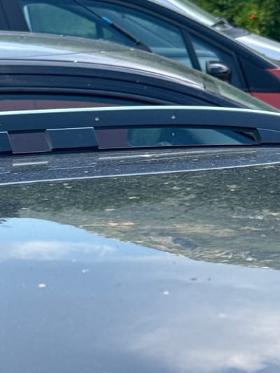 The image is a close-up, vertical shot of the side of a vehicle, possibly a car or van, parked outdoors. The vehicle's dark-colored frame and window are prominent in the foreground. A windshield wiper is visible along the top edge of the frame. Reflected in the vehicle's window is a landscape dominated by a steep, tree-covered mountain slope under a bright, partly cloudy sky. The scene suggests a daytime setting, likely during pleasant weather. The mountain's dense vegetation is a mix of green and lighter hues, indicating a natural, perhaps rural or mountainous area. The sky shows patches of blue and white clouds, with some bright spots that could be the sun reflecting or small, airborne particles. No people are visible. The image's perspective is from slightly above and to the side of the vehicle, focusing on the car's structure and the reflected scenery. The location context of Radanovići, Montenegro, aligns with the mountainous terrain depicted.