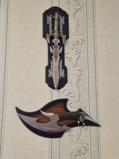 The image displays two decorative knives mounted on a textured, light-colored wall, likely in an indoor setting. The top knife is a sword-like blade with an ornate hilt, mounted on a black plaque with white decorative patterns. Below it, a larger, curved blade knife with a wooden handle wrapped in green cord is affixed to a dark, wood-grain shaped plaque. The wall has a vertical striped pattern and a decorative raised floral motif runs vertically between the two knives. The scene appears to be a still life, with no people or animals present. The lighting suggests a well-lit room, and the overall impression is one of decorative wall art. The location is specified as Новоишимское, Kazakhstan.
