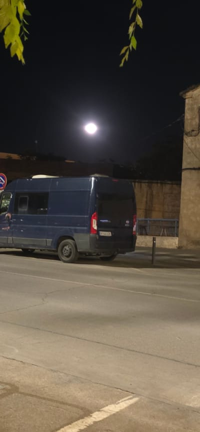 This is a photograph taken at night, likely outdoors, possibly in Huesca, Spain. The main subject is a dark blue van parked on a street. The van has its rear doors closed, and its license plate, which appears to be European, reads "X37 7718". The street surface is visible and appears to be asphalt, with some markings and cracks.
In the background, there is a building or structure made of concrete or stone, with some electrical wiring visible. To the left of the frame, a bright, circular light source, likely the moon, is prominent in the dark night sky. There are also some branches with green leaves in the foreground, suggesting the presence of trees or other vegetation.
The lighting suggests it is nighttime, with artificial light from the moon and possibly streetlights illuminating the scene. The weather appears clear to allow for a visible moon. No people are visible in the photograph. The overall mood is quiet and still, typical of a night scene.