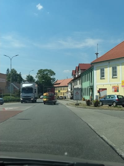 The image shows a street scene in Střelské Hoštice, Czechia. On the right side of the road, there is a row of buildings with red tile roofs and light-colored facades, painted in yellow and green. One building has a sign that says "ZDE V PRODEJI PLYNOVÉ LAHVE". A blue car is parked in front of this building. On the left side of the road, a white truck is driving towards the camera. Behind it, a red truck with a trailer carrying yellow and green objects is also moving away from the camera. A "POZOR" (Attention) sign is visible on the left side of the road, warning of a closure on a road marked with EV.Č. 17210 - 3. The sky is blue with a few white clouds. The road is asphalt.