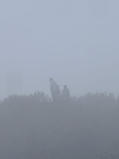 This media file shows a misty scene with three people silhouetted against the fog. Two of the people are standing and appear to be conversing, while the third person is seated. They are positioned in the lower half of the frame, above a dense line of vegetation that is also obscured by the mist. The background is a soft, uniform gray, suggesting a very foggy or overcast day. The overall atmosphere is serene and somewhat mysterious due to the limited visibility. No objects or text are clearly discernible. The time of day is unclear, but the diffused light suggests it could be early morning or late afternoon. The setting appears to be an outdoor natural area. The location context indicates this is Dhundi, India, known for its cool climate and occasional fog, especially in higher altitudes.