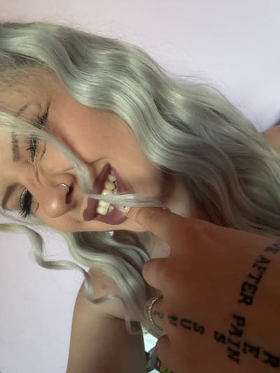 A close-up, low-angle shot captures a woman with light grey, wavy hair. Her head is tilted back, and she is biting on a strand of her hair. Her mouth is open, revealing her teeth, and she is wearing dark lipstick. A ring is visible on her nose. Her right hand is in the frame, with tattoos on the back of her hand that appear to read "SUN S", "RE", and "PAIN AFTER". A ring is on her left ring finger. The background is a plain, light pink surface. The image's specific location in the city of Obruchishte, Bulgaria, is not determinable from the visual information provided.