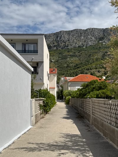 The image shows a narrow, paved alleyway in Dugi Rat, Croatia (latitude: 43.446, longitude: 16.634). The alleyway is flanked by whitewashed buildings, and a stone wall with a wavy pattern on one side. The wall is overgrown with lush green vegetation. In the background, a mountain rises up with a green, forested slope, and several houses with red tiled roofs are visible. The alleyway is empty and leads to the open space at the top of the hill. There are no people or animals visible in the image. The weather is clear and sunny, with blue skies and some white clouds. The alleyway is well-lit and there are shadows from the buildings on the pavement. The image conveys a sense of peace and quiet.