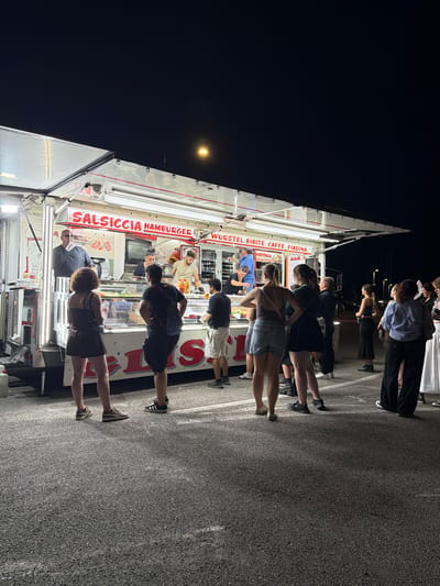 A food truck is parked at night in Segrate, Italy, with people gathered around it. The food truck has "SALSICCIA HAMBURGER WURSTEL BIBITE CAFFE PIADINA" written on its side, indicating it serves sausages, hamburgers, wurstels, drinks, coffee, and piadinas. There are several people in line waiting to order, and some staff members are visible working behind the counter. The scene is illuminated by the bright lights of the food truck and a distant street lamp. The ground is paved asphalt.
