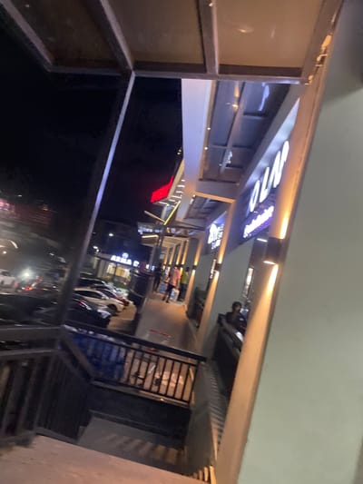 The image depicts an outdoor scene at night, possibly in Guzape, Nigeria, featuring a modern building with a lit facade. A dark, angled structure, likely a walkway or stairway, dominates the foreground, with a black metal railing. Beyond this, a paved walkway is visible, lined with parked cars and illuminated by building lights. Several individuals are present, some walking along the pathway and others seated, possibly at an establishment. Neon signs, including one with large white lettering that appears to spell "OLAF", are affixed to the building. The overall atmosphere suggests a commercial or entertainment area, active in the evening.