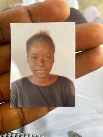 A headshot photograph of a young Black woman is held between the thumb and forefinger of a person's hand, obscuring the lower left and lower right corners of the photograph. The woman in the photograph has dark skin, short, dark, curly hair, and is wearing a dark crew-neck shirt. The background of the photograph is a plain, light color. The hand holding the photograph has dark skin and is resting on a white surface. The image was taken in Jimeta, Nigeria.