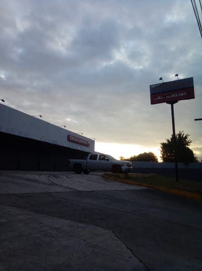 The image shows a silver pickup truck parked outside a commercial building with a sign that reads "COAVENCHO". The building is a light gray color and appears to be a warehouse or retail space. A large sign on a pole is visible in the background with the letters "CHOA." The sky is overcast with a hint of sunrise or sunset on the horizon. The foreground shows a paved area leading up to the building. The location is Parroquia Guatire, Venezuela.
