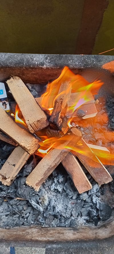 A close-up, top-down view captures a small bonfire burning in a metal brazier. Several pieces of light-colored, unfinished wood are arranged in a crisscross pattern, with one piece at the bottom left showing a partial image of a woman and text, possibly from a magazine or packaging. Bright orange and yellow flames lick upwards, obscuring some of the wood and ash below. The brazier itself is dark gray and appears to be made of stone or concrete. The background above the brazier is a textured, dark olive-green surface. The scene is set outdoors, with the context of the city of Iskrets, Bulgaria implied.