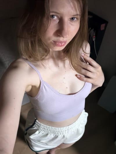 A young woman with light brown hair and light blue eyes is lying down, looking up at the camera. She is wearing a light purple tank top and white shorts. Her left arm is bent, with her hand resting on her chest. The background is dark and appears to be an indoor setting. There are some objects in the background, including what looks like a computer monitor. The lighting suggests it is nighttime or a dimly lit room. The woman has a neutral expression. No visible text is present in the image. The location context is Dongducheon-si, South Korea.