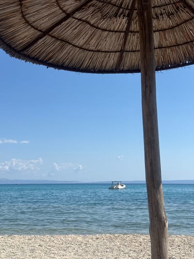 The image depicts a serene beach scene in the Municipal Unit of Pallini, Greece. In the foreground, a weathered wooden pole supports a straw umbrella, its textured surface suggesting age and exposure to the elements. Beyond the pole, a shoreline composed of small, light-colored pebbles stretches across the bottom of the frame. The calm, turquoise sea occupies the middle ground, its surface gently rippled. In the distance, a small white motorboat with an open cabin is anchored or drifting on the water. The horizon is marked by a faint, hazy line of land, possibly an island or coastline. Above, a clear, bright blue sky dominates the upper portion of the image, with a few wispy white clouds scattered near the horizon. The overall atmosphere is one of tranquility and a typical summer day at the beach.