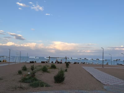 The image depicts a beach scene in Çolpon-Ata, Kyrgyzstan, along the shores of Issyk-Kul Lake. The sky is a pale blue with scattered, fluffy white clouds, tinged with pink and orange from what appears to be sunset. The water is a deep blue, calm, and stretches to the horizon.

The sandy beach is lined with white canopies providing shade for lounge chairs. Several people are visible relaxing under these canopies. In the foreground, a paved walkway with a red and white checkered pattern leads away from the viewer towards the beach. Small, green bushes are sparsely planted on the sand. To the left of the walkway, a lifeguard tower with a ladder stands near the water's edge. Streetlights are positioned along the beach.

The overall atmosphere is one of tranquility and relaxation, characteristic of a resort town.