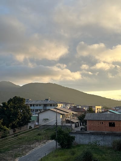 The image depicts a cityscape in Florianópolis, Brazil, situated against a mountain backdrop under a partly cloudy sky. The foreground reveals a sidewalk with a fence. Beyond that, there is a row of buildings, varying in height and construction material (brick, concrete). Overhead power lines run parallel to the street. In the background, a mountain rises steeply, partially obscured by clouds. The lighting suggests either early morning or late afternoon.