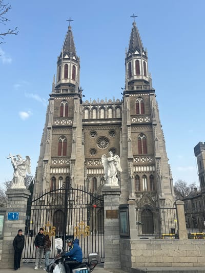 The image depicts the Hongjialou Cathedral in Licheng District, Jinan, China, viewed from a low angle on a clear day. The prominent structure is a large Gothic-revival style cathedral featuring two symmetrical spired bell towers, each topped with a cross. The building's facade is constructed from grey stone and reddish-brown brick, showcasing multiple levels of arched windows, buttresses, and decorative stone carvings. A large circular rose window is centered on the upper facade.

In the foreground, a black wrought-iron gate, adorned with the gilded letters "JHS," blocks access to the cathedral grounds. The gate is flanked by two large white marble statues of angels standing on stone pedestals. The angel on the left holds a cross, while the angel on the right holds a wreath or crown. A blue street sign displaying the number "1" is affixed to the left pedestal.

On the paved area in front of the gate, four individuals are present. To the left of the gate, three people stand near the left pedestal: a male in a dark jacket, a male in a black hooded sweatshirt, and a female in a plaid top, conversing. To the right, a male in a blue jacket and a dark helmet operates a black and blue scooter, facing left. The scooter is equipped with a front basket holding a white package and a rear storage box. Bare tree branches are visible against the clear blue sky on both sides of the cathedral, and a smaller, older stone building is partially visible to the far right.