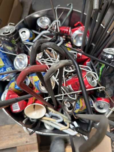 The image shows a close-up, overhead view of a metal bin filled with various metal items and crushed aluminum cans. The bin appears to be a recycling bin. Among the items are several red and black cans, some with logos that are partially visible, including a Monster Energy can. There are also numerous bent metal rods and wires, some of which are dark gray and others a lighter gray or reddish-brown. The context suggests this is a scene from Mol, Belgium, likely in an industrial or workshop setting where metal waste is collected for recycling. The lighting is dim, and the focus is on the contents of the bin, with the background out of focus, showing hints of cardboard boxes and wooden pallets, reinforcing the idea of a storage or disposal area. There are no people visible in the image.