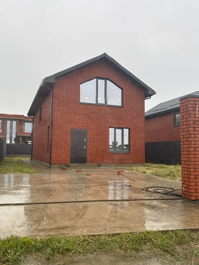 The image depicts a newly constructed brick house, likely in the village of Starobzhegokay, Russia, as the user specified the location. The house features a prominent triangular window on its gable end and a standard rectangular window beside a black entrance door. The exterior is made of dark red bricks with gray window frames and roof trim. A wet concrete walkway runs alongside the house, with grass growing along the edge. A black garden hose and various tools are scattered on the walkway, indicating recent or ongoing construction activities. The sky is overcast, suggesting a recent rainfall event. No people or interactions are visible in the image. The scene overall suggests a new construction site in wet weather conditions.