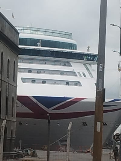 The image displays a large, white cruise ship docked in the Port of Montevideo, Uruguay, under an overcast sky. The ship's bow features a prominent red, white, and blue stylized geometric design. Multiple decks are visible, including a glass-enclosed upper observation lounge and an open deck where one individual in a light-colored top is observed. The bridge area is also distinct. To the left, a portion of a multi-story, grey stone or concrete building with arched window openings is visible. A dark utility pole with an attached streetlight and a visible "40" sign stands vertically on the right, partially obscuring the ship. In the lower foreground, a metal fence is present, and indistinct objects consistent with port operations are visible behind it. A faintly discernible individual walks in the lower right corner of the frame.