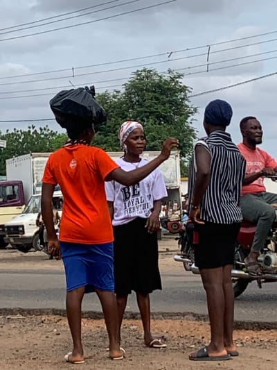An outdoor scene in the city of Shendam, Nigeria, captures four individuals on an overcast day. In the foreground, a person with dark skin, wearing an orange t-shirt and blue shorts, stands facing right, balancing a black plastic bag on their head. They wear white flip-flops. To their right, an adult female with dark skin wears a white headwrap, a white t-shirt with partially visible text "BE LOYAL EVERYTHI," a black skirt, and white flip-flops. She stands facing left, gesturing with her right hand, index finger extended, towards the person with the bag on their head. Immediately to her right, a third person with dark skin stands with their back partially to the camera, wearing a dark blue headwrap, a black and white striped shirt, black shorts, and dark slide sandals. Further right, an adult male with dark skin is seated on a red motorcycle, facing right and slightly away from the other individuals. He wears a red t-shirt (with "COWBOYS" partially visible), dark trousers, and dark sandals. The ground beneath the individuals is unpaved dirt and gravel, transitioning to a paved road surface in the mid-ground. Various vehicles, including a white car and a white truck, are visible on the road and in the background. Green foliage and indistinct structures are present in the far background. Multiple power lines span across the cloudy, light grey sky.