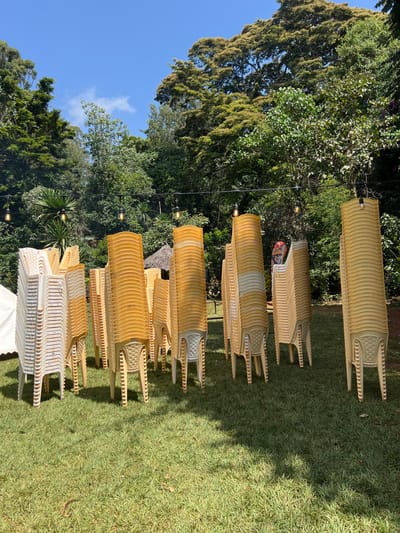 The image, oriented correctly, displays an outdoor setting featuring numerous stacks of plastic chairs on a manicured lawn. The scene is dominated by vibrant green grass in the foreground and midground, with dense, lush green trees and foliage forming a natural backdrop. A clear blue sky is visible in the upper left portion of the image.
The main subjects are the chairs, which are neatly stacked in several columns. Most of the chairs are a light yellowish-beige color, while a stack on the far right includes white chairs. There are at least seven distinct stacks of chairs visible. String lights with visible bulbs are strung across the upper part of the image, extending from left to right, indicating preparations for an event or adding to the aesthetic lighting of the venue.
The setting appears to be a garden or an event space, likely in Mururiini, Kenya, given the context. The bright sunlight suggests it is daytime, possibly late morning or early afternoon, with crisp shadows cast on the grass by the chair stacks. The weather is clear and sunny. There are no people visible, and no discernible activity other than the static arrangement of the chairs, suggesting either preparation for an upcoming gathering or the cleanup after one. No visible text or explicit emotional cues are present. The overall impression is one of an organized outdoor event area amidst natural beauty.