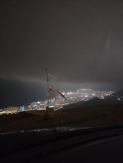 The image captures a panoramic nighttime view of a coastal city, identified as Alto Hospicio, Chile, illuminated by a myriad of artificial lights. The scene is presented from an elevated perspective, looking down and across the sprawling urban landscape.

In the foreground, on the bottom right, there is a dark, rough-textured surface, possibly a cliff edge or a concrete structure. From this edge, a tall, slender pole extends diagonally upwards and towards the left, with several pieces of tattered fabric or flags tied to its top. These fabrics appear to be in shades of white, red, and yellow, fluttering slightly.

Beyond this foreground and pole, the brightly lit city stretches horizontally across the mid-ground of the image, outlining what appears to be a coastline or the edge of a large bay. The individual lights of buildings and streets create a dense, sparkling pattern against the darkness. Below the city lights, a dark expanse of water is visible, reflecting some of the city's glow as indistinct streaks of light.

The sky above the city and water is dark and appears overcast or foggy, with a subtle, diffuse glow emanating from the city's light pollution reflecting off the atmospheric conditions. There are no discernible stars or moon. The overall impression is one of a vast, tranquil night scene. No visible text is present in the image.