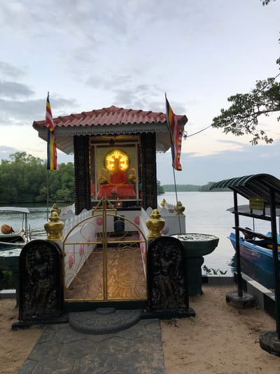The image depicts a Buddhist shrine situated by a body of water in Habaraduwa, Sri Lanka. A statue of Buddha, illuminated by a golden halo, is housed within a small, ornate structure topped with a red tiled roof. Two Buddhist flags are positioned to the left of the shrine. In the foreground, a golden gate and decorative stone carvings flank the entrance. To the right, a blue boat is docked next to a shaded waiting area. The background features lush green trees and a calm expanse of water, indicative of a natural or semi-natural environment. The sky is overcast. There are no people visible in the image.