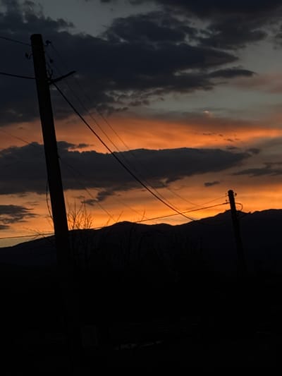 This is a photograph taken in Mikrevo, Bulgaria, depicting a sunset scene. The sky is a dramatic display of oranges and dark grays, with clouds creating a textured backdrop. In the foreground, two silhouetted utility poles stand starkly against the vibrant sky. Wires crisscross between the poles, adding a linear element to the composition. Below the sky, a dark, silhouetted mountain range forms the horizon line. The lower portion of the image is almost entirely black, obscuring any ground-level details. The overall impression is one of a rural, twilight landscape.